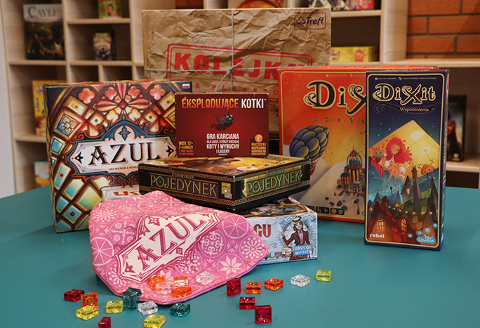 Na stole w bibliotece znajdują się postawione gry planszowe, takie jak: Azul, Eksplodujące kotki, Pojedynek, Dixit. Dodatkowo przed grami rozsypane kwadratowe, kolorowe żetony do gry w Azula.