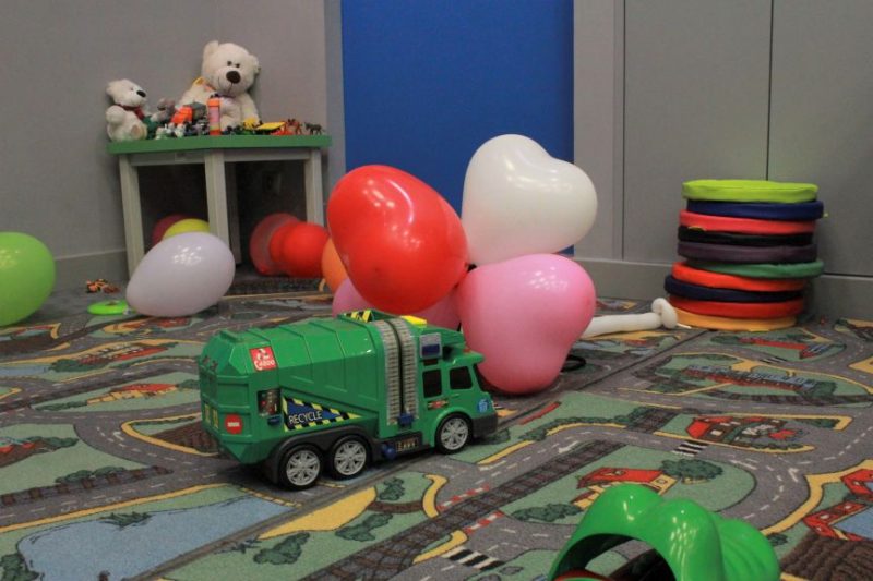 kacik dla dzieci, na dywanie zabawkowe auto, za nim pęk balonów, pod ścianą stoi stolik, na nim miś